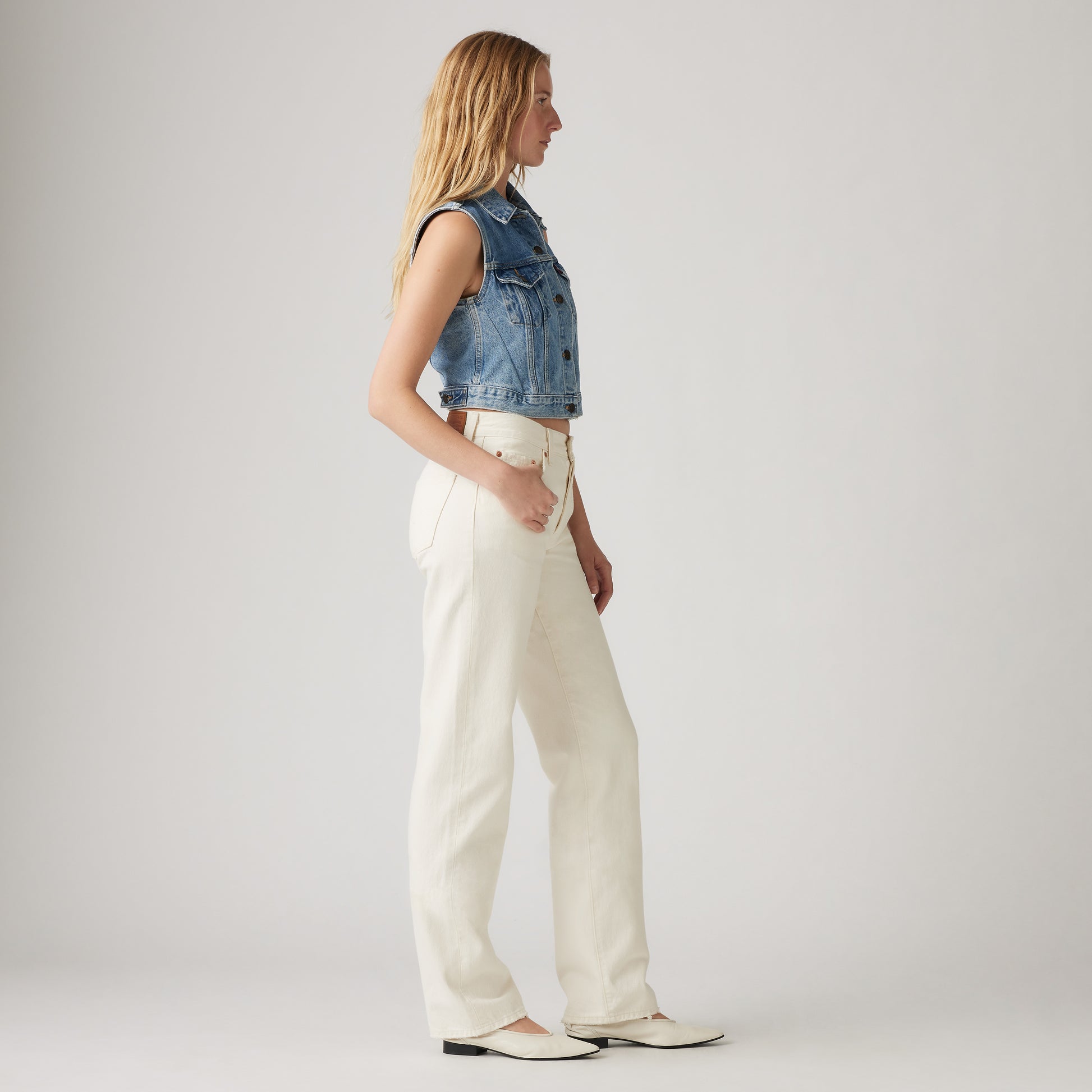 A woman stands in profile against a plain light background, wearing mid-rise white Levis 501 90s jeans, a sleeveless denim vest, and white shoes.