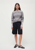 An individual stands against a plain background wearing a gray sweater with handcrafted horn buttons, Aiayu's Willy Shorts in black, and black sandals.