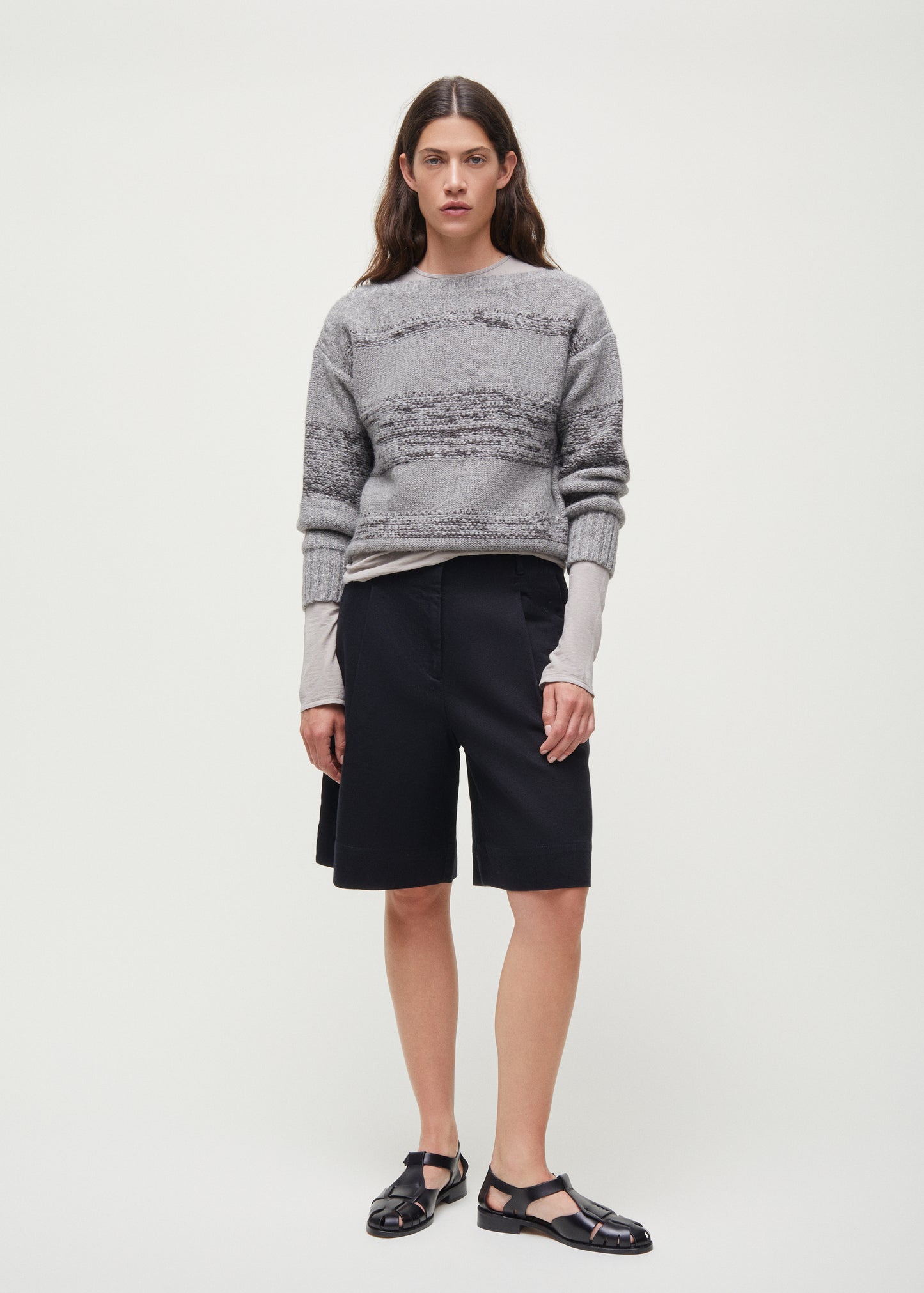 An individual stands against a plain background wearing a gray sweater with handcrafted horn buttons, Aiayu's Willy Shorts in black, and black sandals.