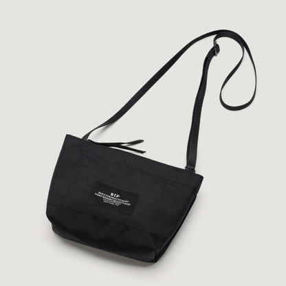 A black Zipper Pouch Mini with a long strap and a white label on the front against a white background from Bags in Progress.
