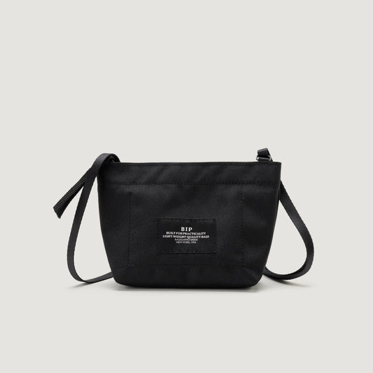 A black crossbody bag with a front Zipper Pouch Mini and adjustable strap, featuring a Bags in Progress label with text in the center.