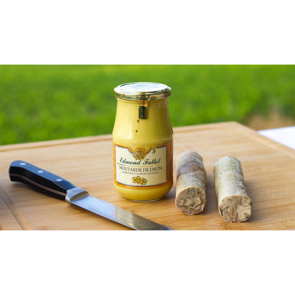 A jar of Edmund Fallot Dijon Mustards from Gourmet Food Solutions, Inc., two sausages, and a kitchen knife displayed on a wooden cutting board, with a green outdoor background.