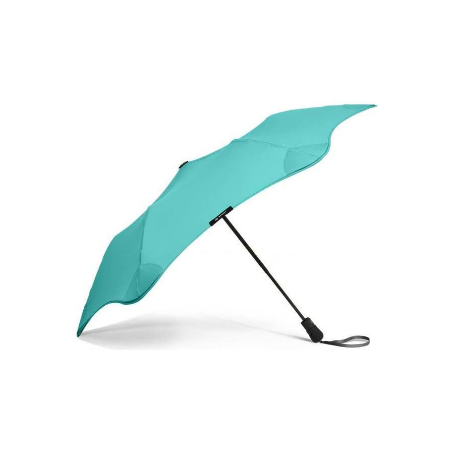 The Blunt Mini Metro Umbrella, shown open and lying on its side against a white background, features a turquoise canopy with a curved, asymmetrical shape and a black handle.