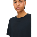 A person with short hair is wearing the Clara Base Tee by Rohnisch, a plain black T-shirt made from recycled polyester, featuring subtle text on the chest against a black background.