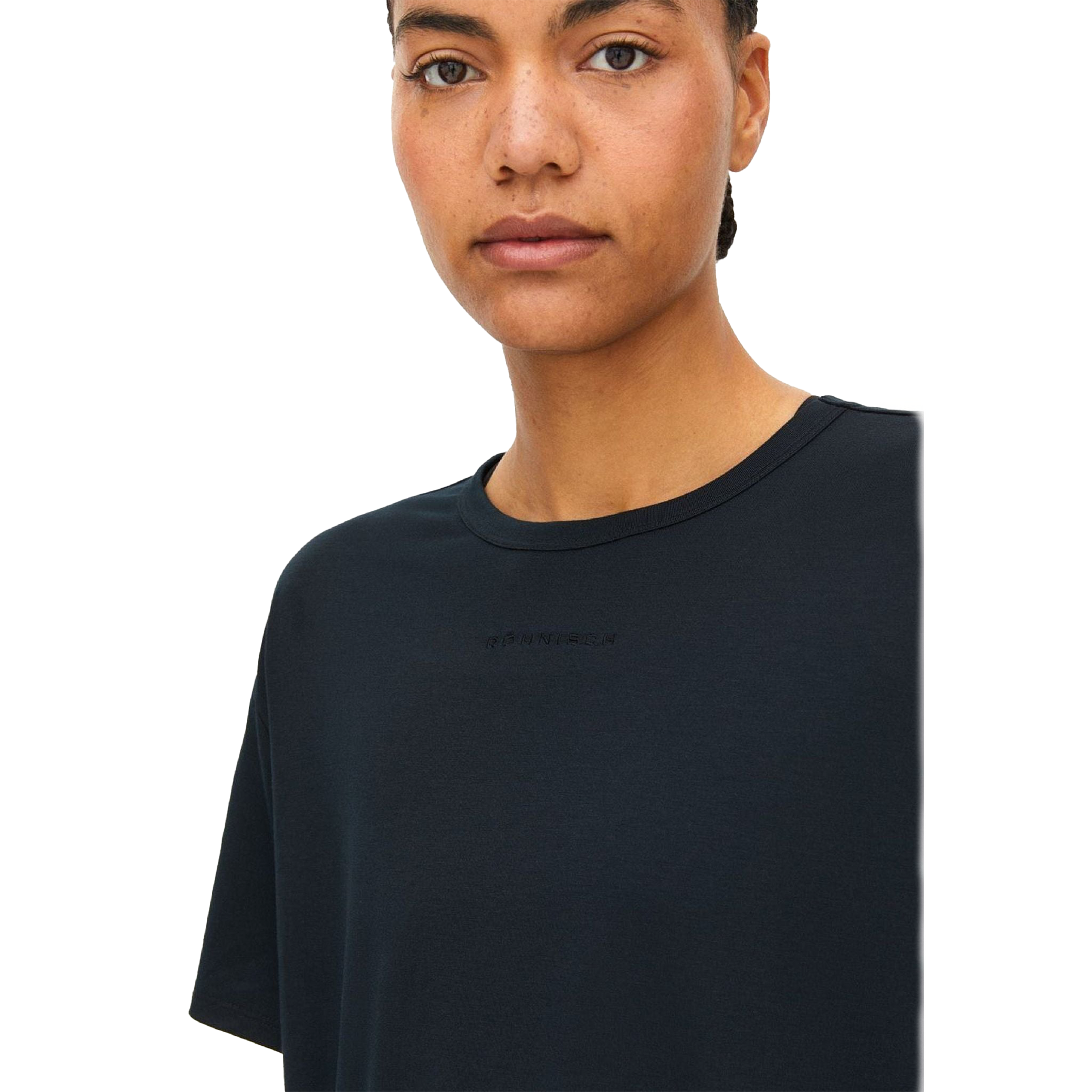 A person with short hair is wearing the Clara Base Tee by Rohnisch, a plain black T-shirt made from recycled polyester, featuring subtle text on the chest against a black background.