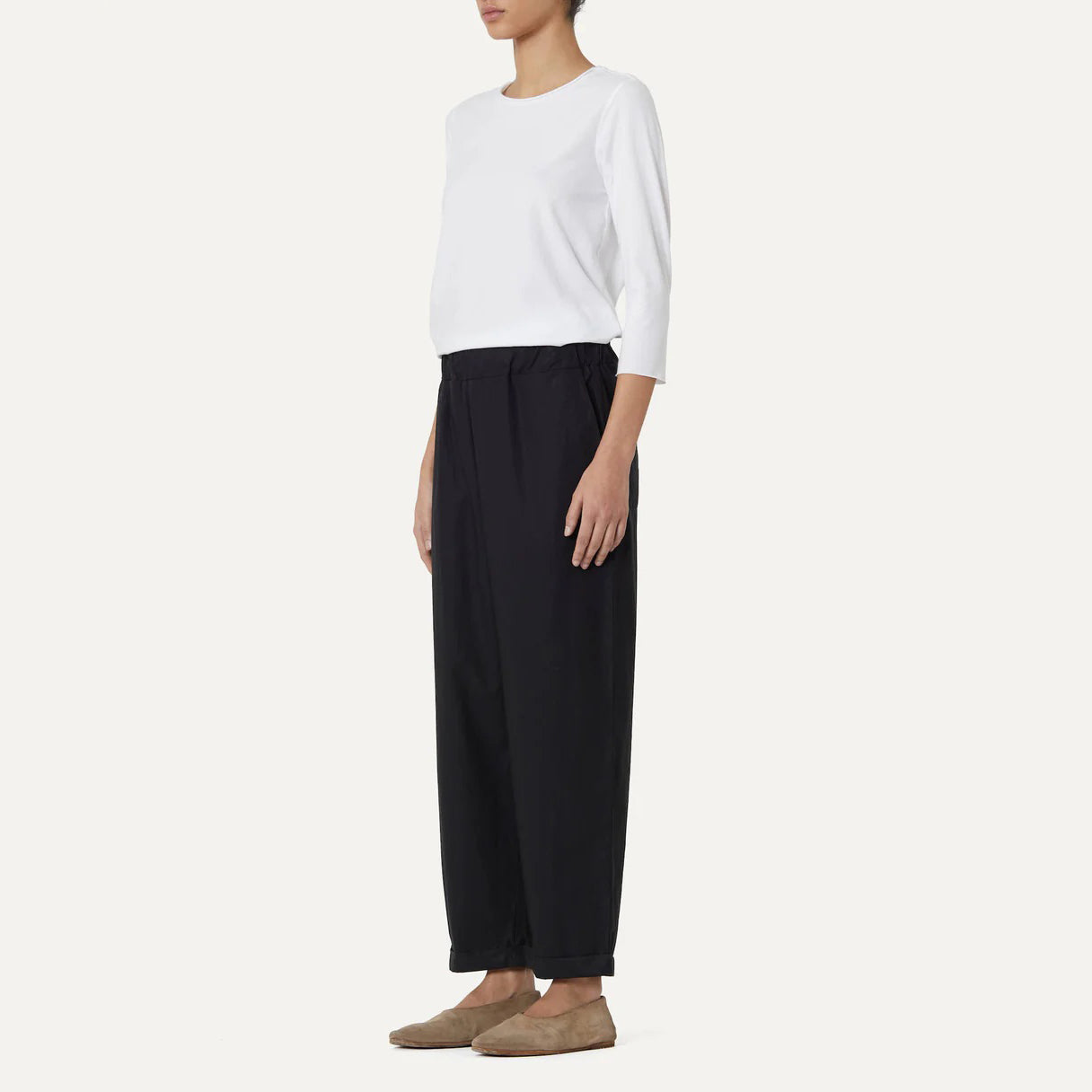 A person in a white top, Panta Vela Pant by Labo Art in black cotton poplin with an elastic waistband, and brown shoes stands against a plain background.