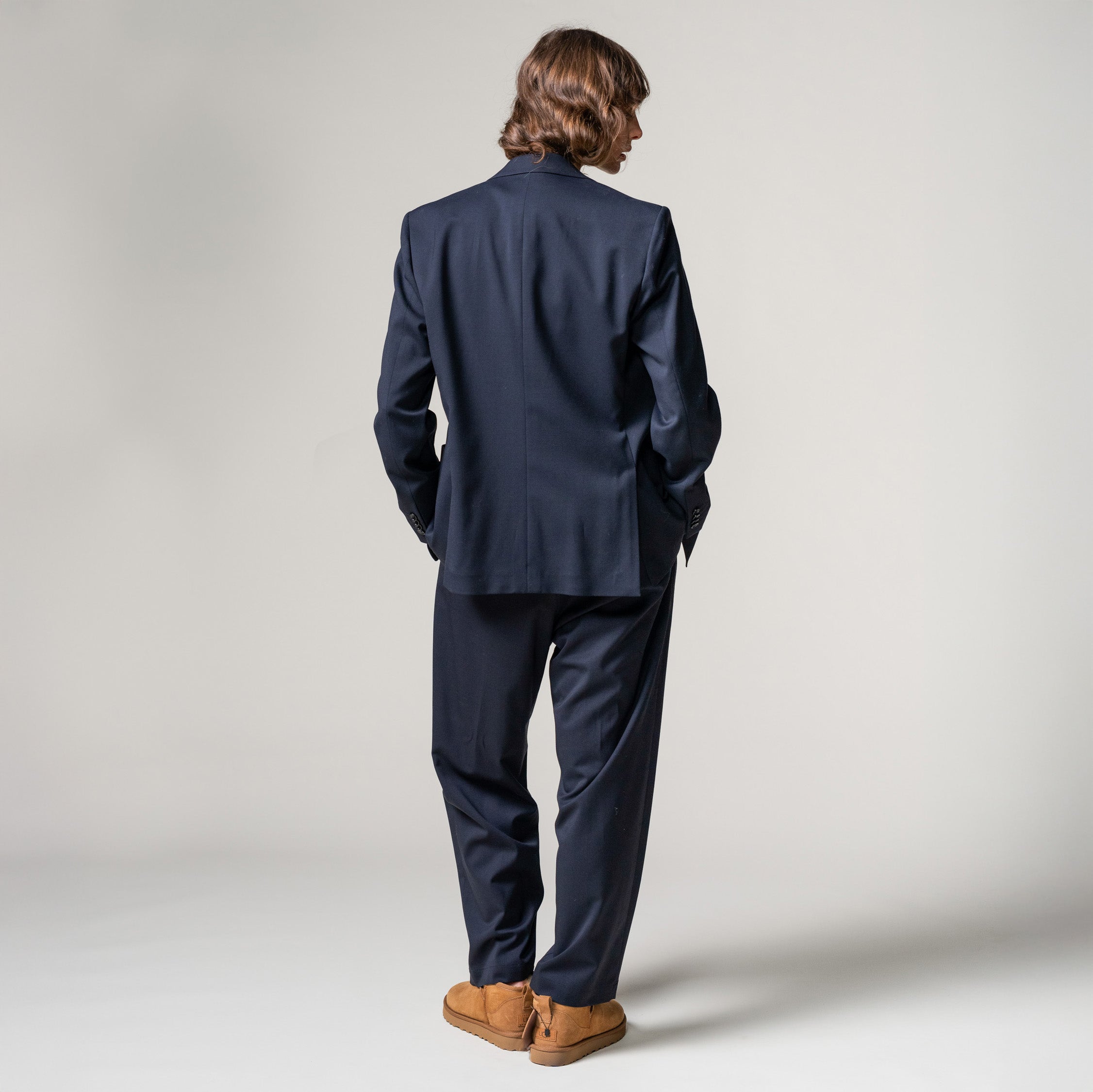 A person with long hair stands facing away against a plain background, wearing a navy Double Breast Blazer Tropical Wool by Seven Gauge and brown shoes.