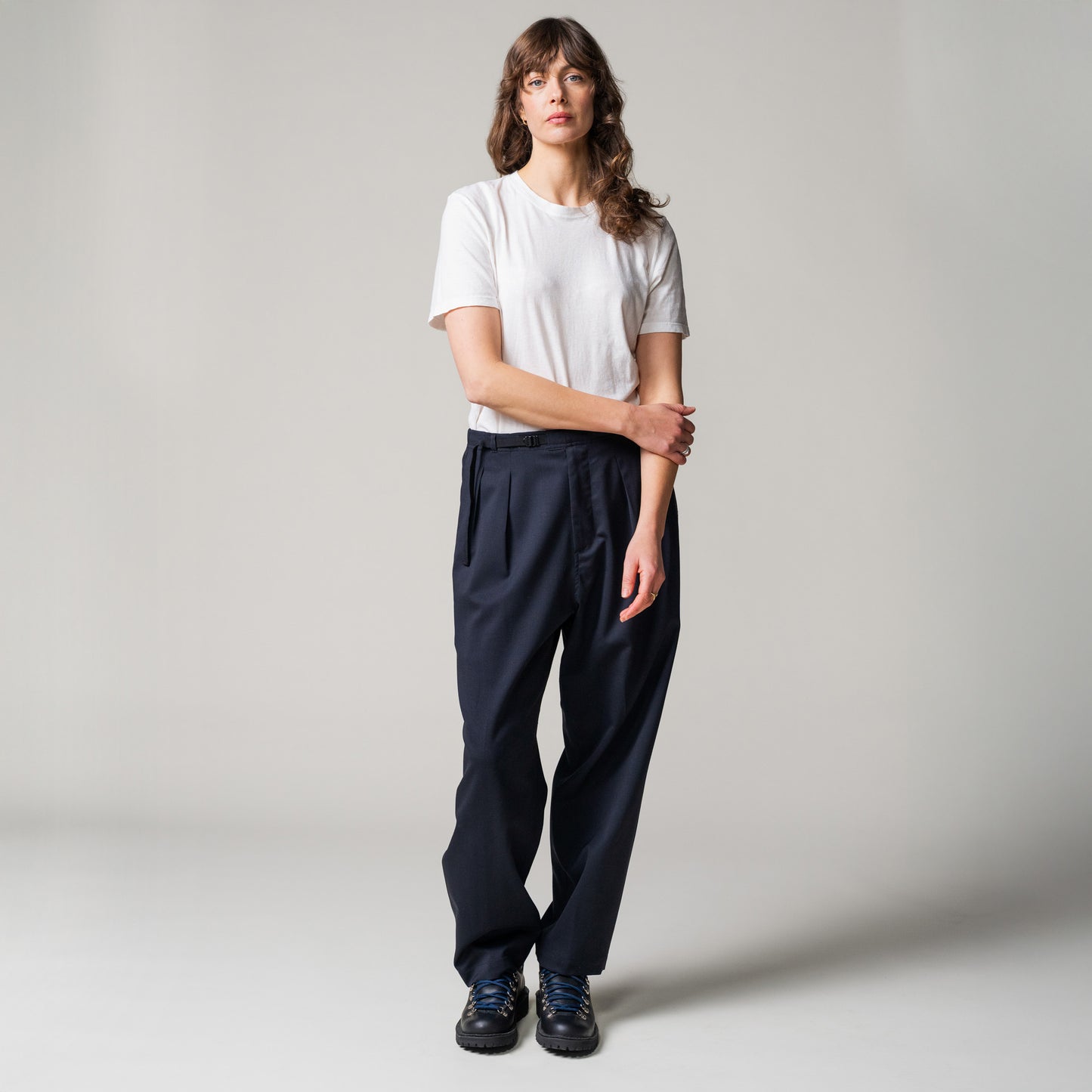 A person stands against a plain background, wearing a white t-shirt, black shoes, and Seven Gauge's blu Trouser Tropical Wool with hands clasped in front; the classic tailoring exudes elegance and sophistication.