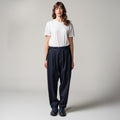 Person standing against a plain background, wearing a white t-shirt, dark blue pleated tropical wool trousers by Seven Gauge made in Italy, and black shoes.