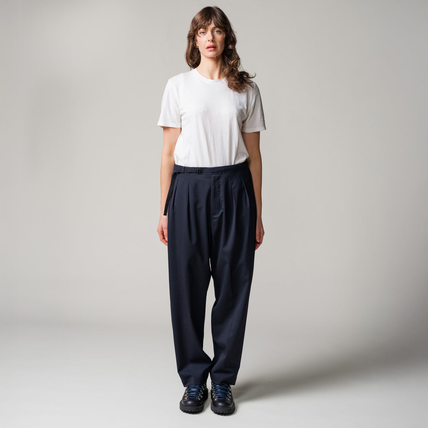 Person standing against a plain background, wearing a white t-shirt, dark blue pleated tropical wool trousers by Seven Gauge made in Italy, and black shoes.