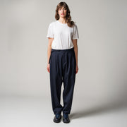 Person standing against a plain background, wearing a white t-shirt, dark blue pleated tropical wool trousers by Seven Gauge made in Italy, and black shoes.