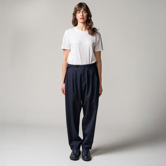 Person standing against a plain background, wearing a white t-shirt, dark blue pleated tropical wool trousers by Seven Gauge made in Italy, and black shoes.