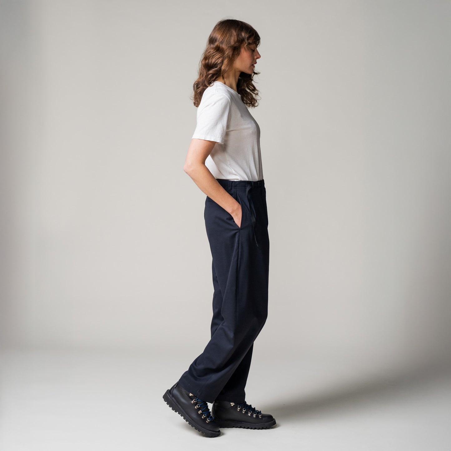 A person stands sideways with hands in pockets, wearing a white t-shirt and classic black Seven Gauge "Trouser Tropical Wool, Blu" pants paired with black shoes against a plain background.