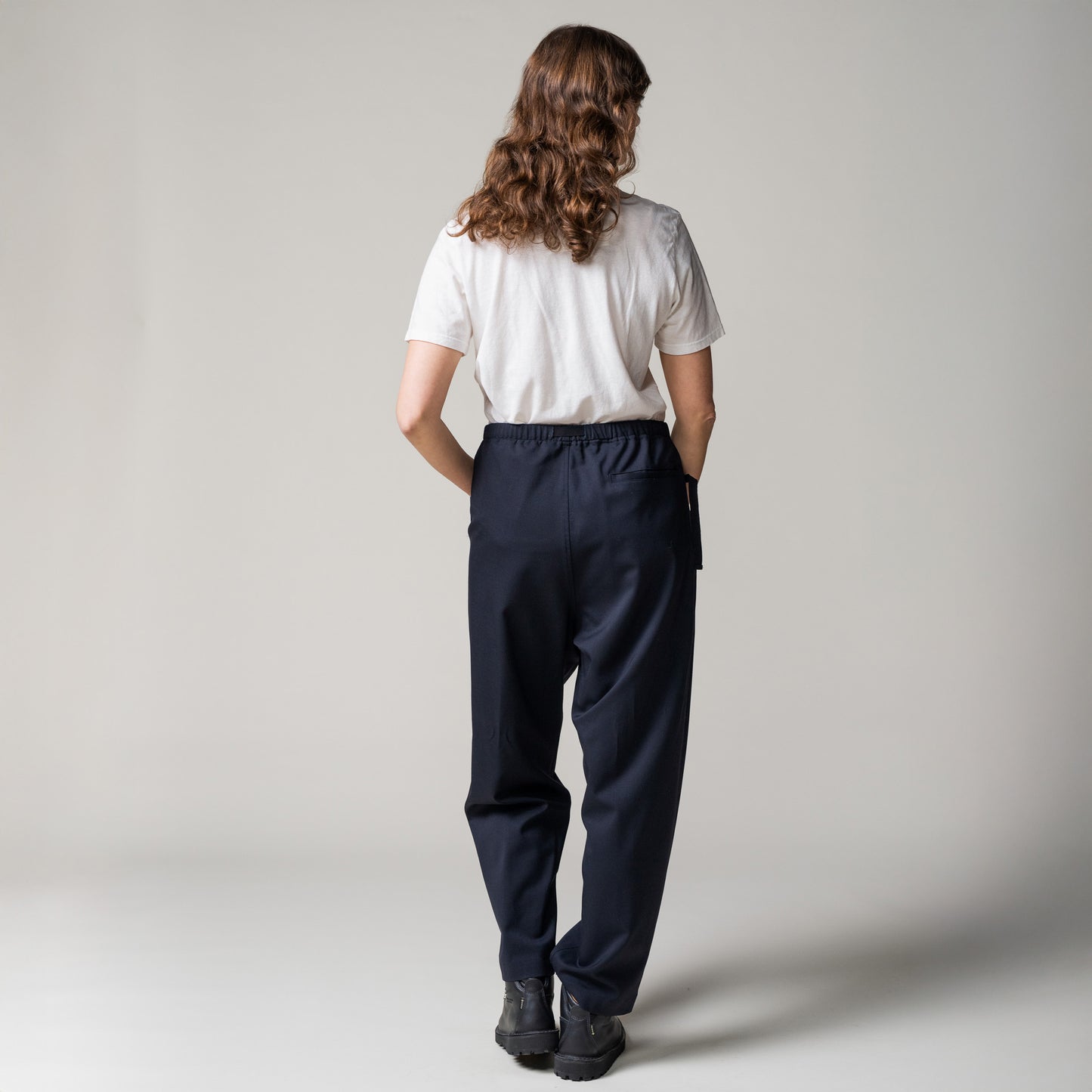 Clad in a white shirt and expertly tailored "Trouser Tropical Wool, Blu" by Seven Gauge, a person with long hair faces away against a neutral backdrop. Their black shoes complement the look, embodying classic tailoring elegance.