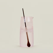 The Essential Glassware - Pitcher by Hawkins New York is a tall, translucent pink borosilicate glass piece with retro charm, shown filled with ice water and a metal spoon against a simple light background.