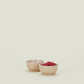 On a light surface, two blush ceramic bowls from Hawkins New York sit; one empty and the other filled with raspberries. These Essential Extra Small Bowls are microwave and dishwasher safe, offering both practicality and style for any kitchen.