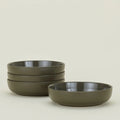 The Essential Low Bowl (Set of 4) by Hawkins New York features four olive-green stoneware bowls, stacked in two piles on a plain light background. Microwave and dishwasher safe, they combine simple elegance with everyday convenience.