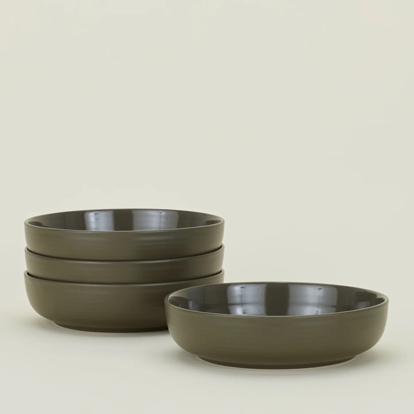The Essential Low Bowl (Set of 4) by Hawkins New York features four olive-green stoneware bowls, stacked in two piles on a plain light background. Microwave and dishwasher safe, they combine simple elegance with everyday convenience.