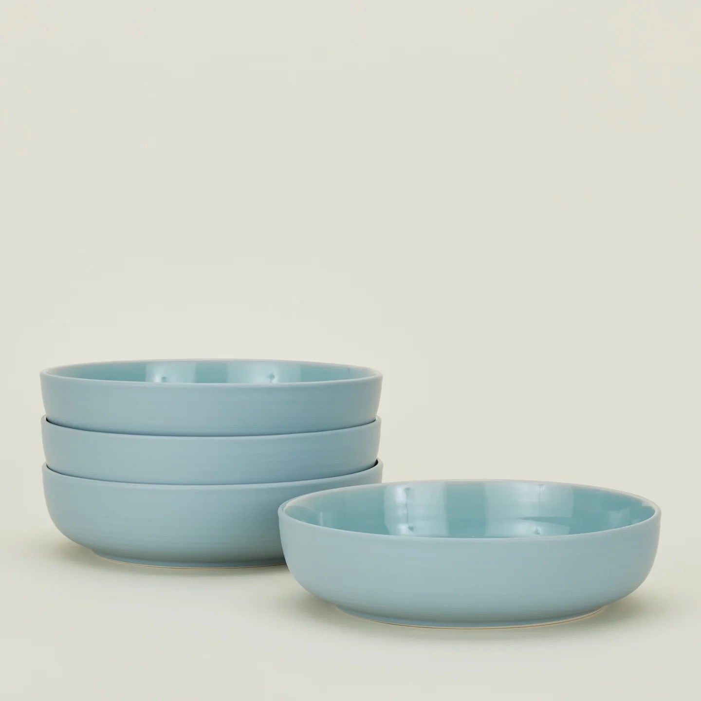 Four Hawkins New York Essential Low Bowls in light blue stoneware are stacked on the left with one in front, all on a cream background. This set of four is microwave safe and dishwasher safe.