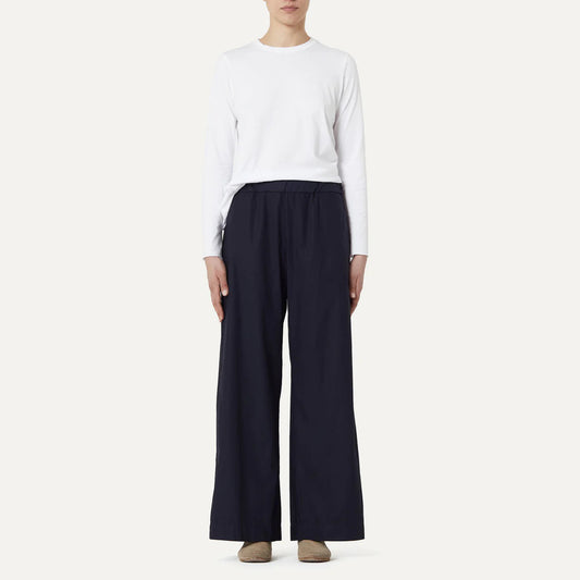 A person confidently stands against a plain background wearing a crisp cotton poplin white long-sleeve shirt, Panta Diana Pant Clara in dark color with subtle patch pockets by Labo Art, and brown shoes.