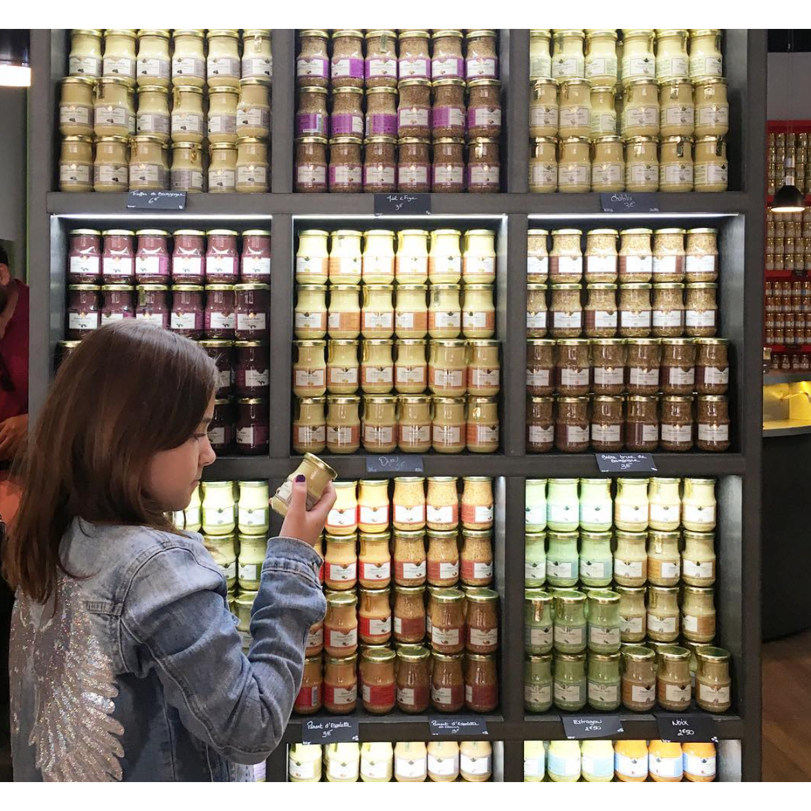 A child in a denim jacket examines a jar of Edmund Fallot Dijon Mustards from Gourmet Food Solutions, Inc. in front of a display filled with jars of various preserves on shelves.