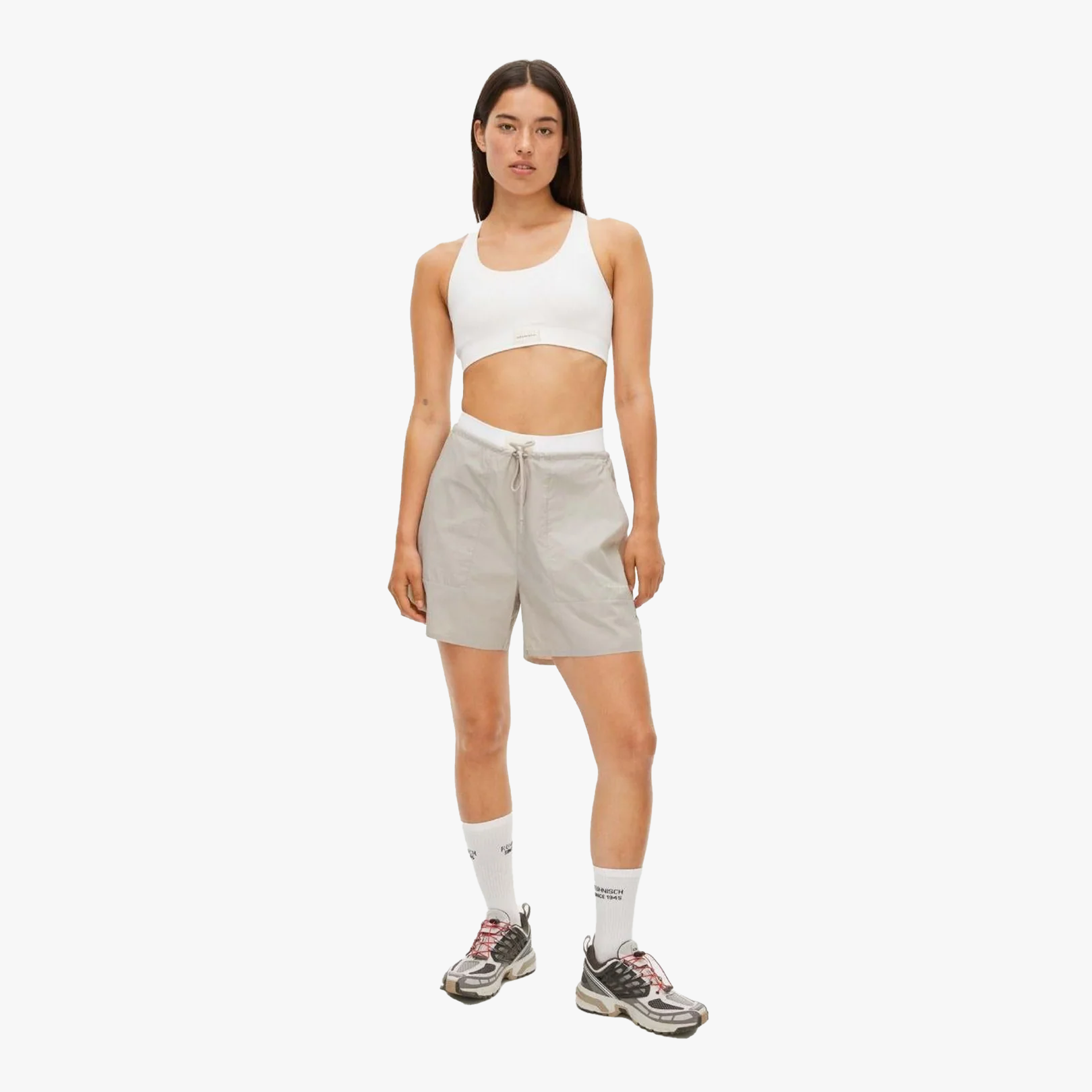 A woman stands against a white background wearing a white sports bra, light gray Rohnisch Frankie Wind Shorts made from polyamide, white socks, and gray sneakers.