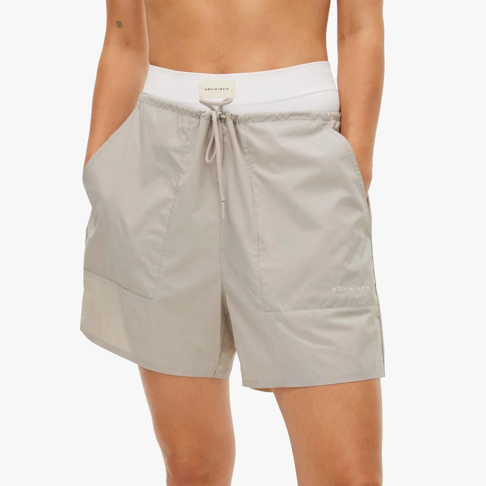 Person wears light gray Rohnisch Frankie Wind Shorts, featuring large front pockets and a white elastic waistband, made from lightweight polyamide for comfort and style.