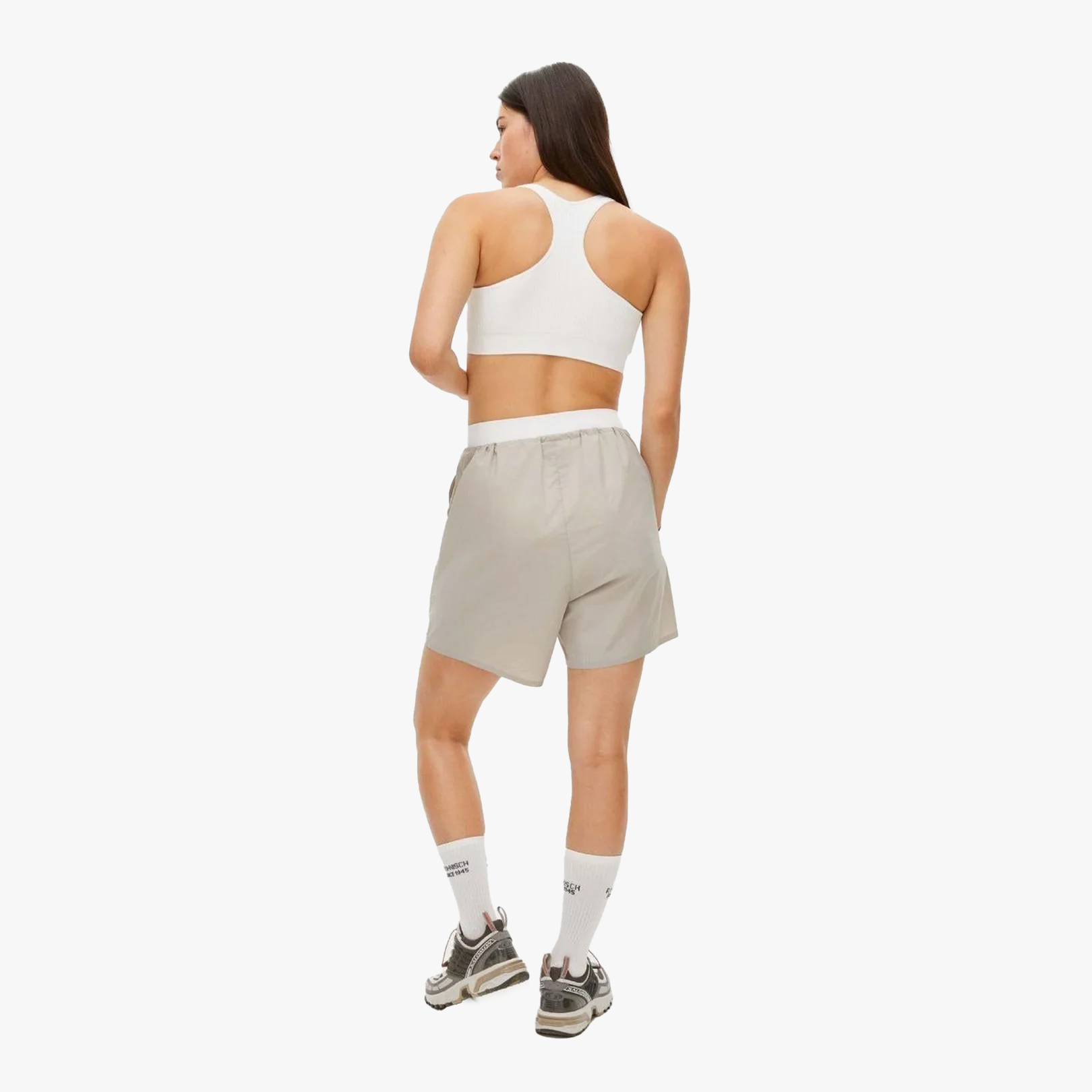 A woman in a white sports bra, Rohnisch Frankie Wind Shorts, white socks, and sneakers stands facing away and looks over her shoulder against a plain white background.