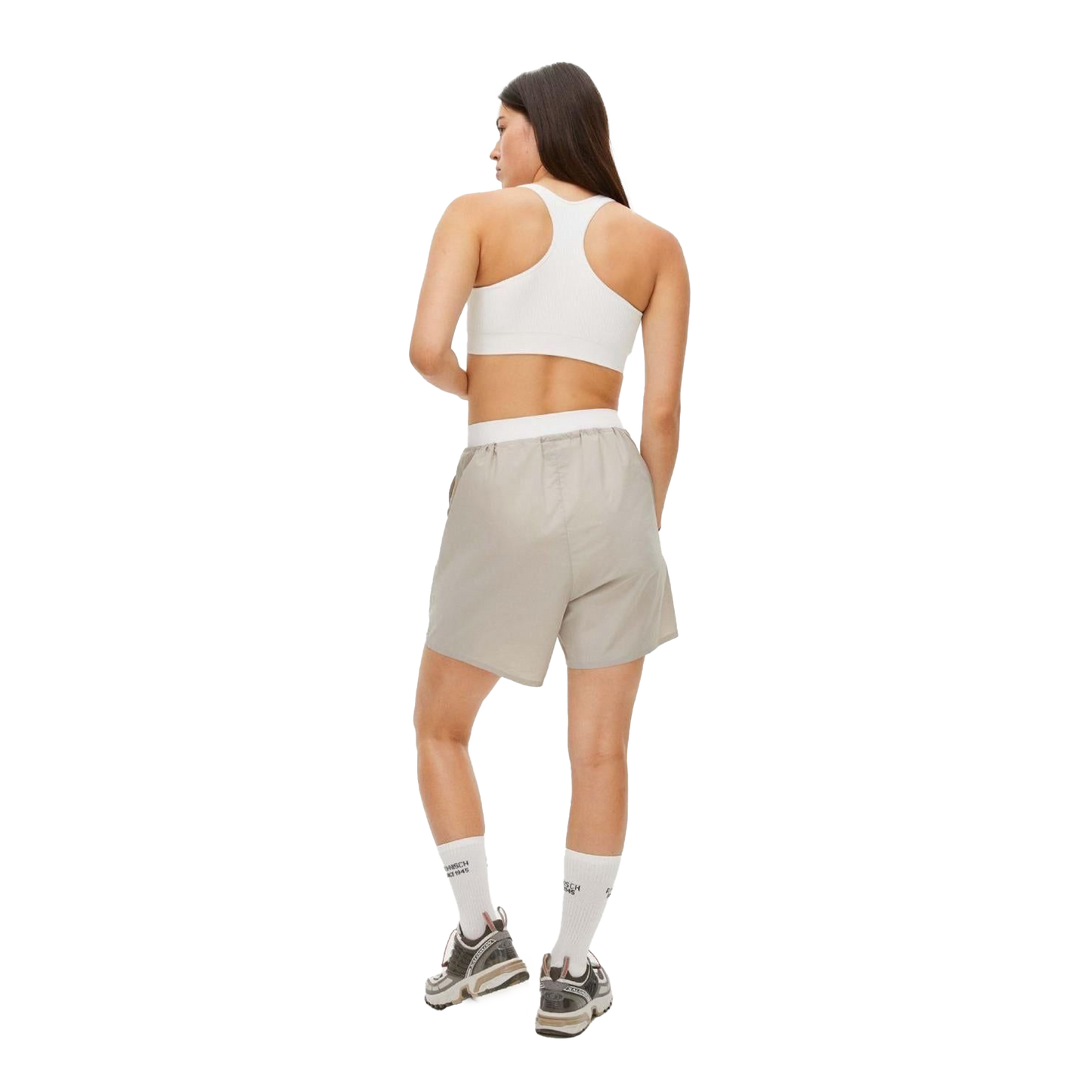 A person faces away, wearing a white sports bra and beige Frankie Wind Shorts by Rohnisch, featuring the logo. They pair them with white socks and gray sneakers.
