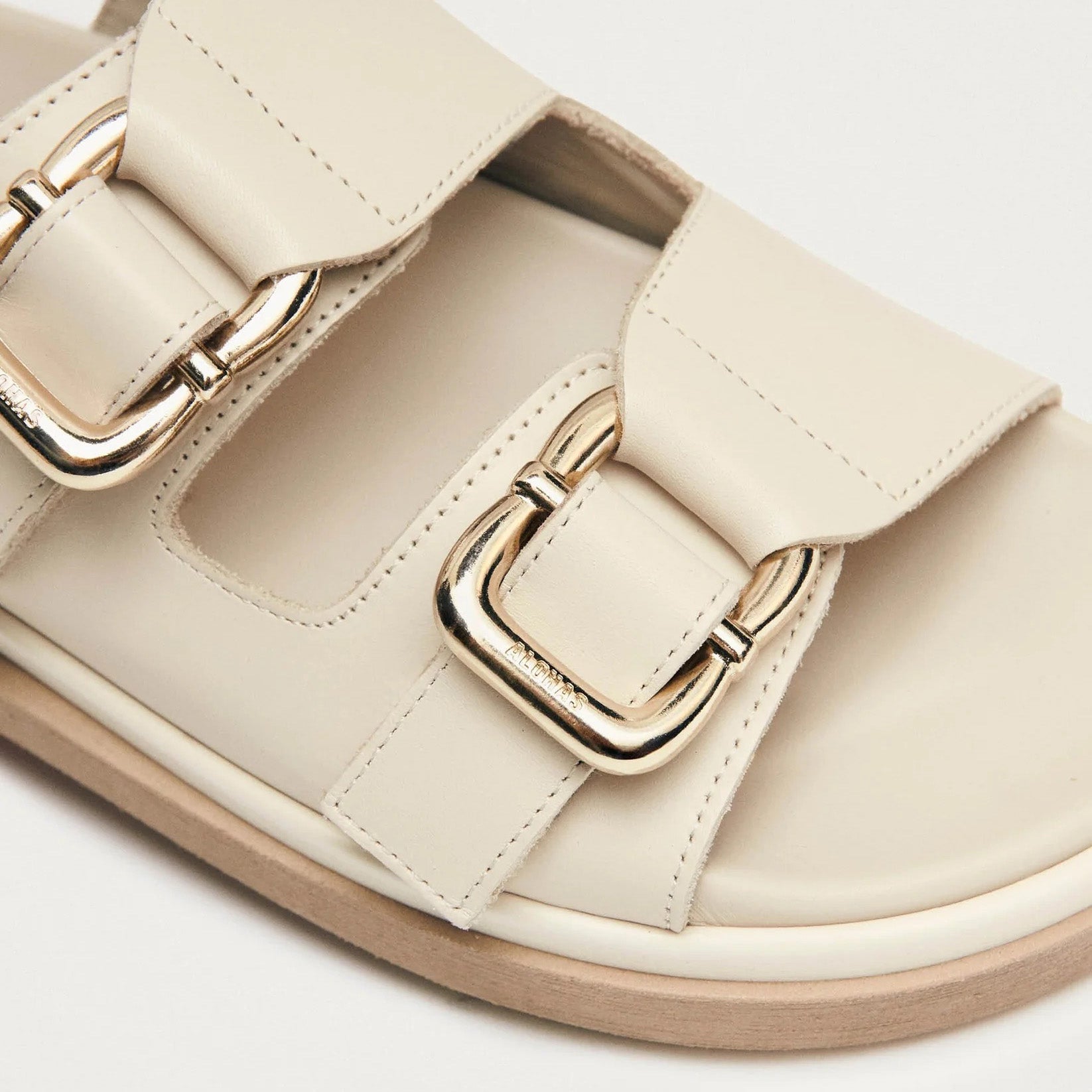Close-up of Alohas Harper Sandal in cream, featuring two wide buckle straps with shiny gold buckles and a light brown sole.