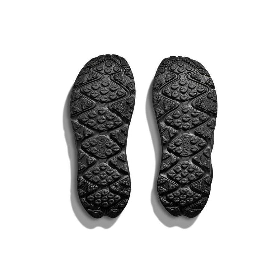 The image displays the textured soles of Hoka's U Ora Primo black slip-on comfort shoes, placed side by side on a white background.