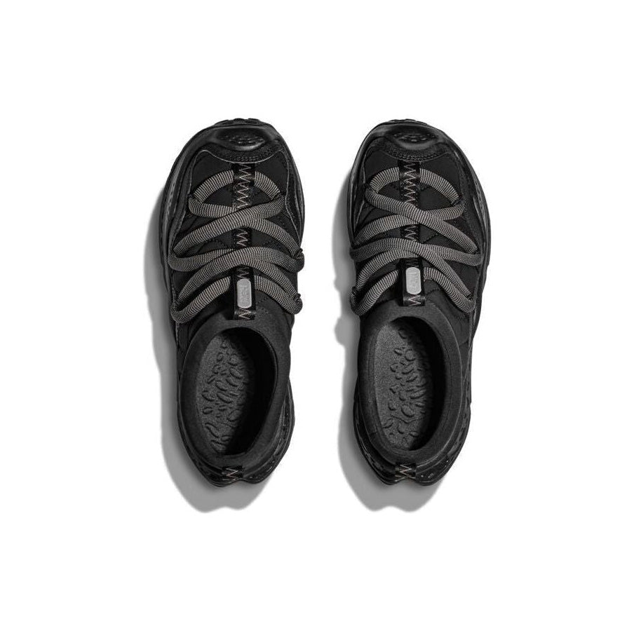 The Hoka U Ora Primo sneakers, in black, feature gray-green crisscross laces and insulated puff uppers for slip-on comfort. Set against a plain white background, these shoes offer style and recovery with every step.