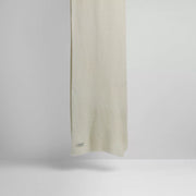 A plain, light-colored Le Bonnet Scarves, Snow lambswool scarf hangs against a neutral background highlighting its biodegradable properties with a small tag near the edge.