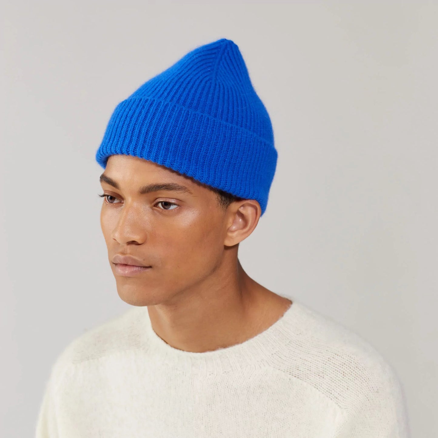 The person wears a Le Bonnet Beanie in Royal Azure, paired with a light sweater, gazing thoughtfully to the side against a plain backdrop.