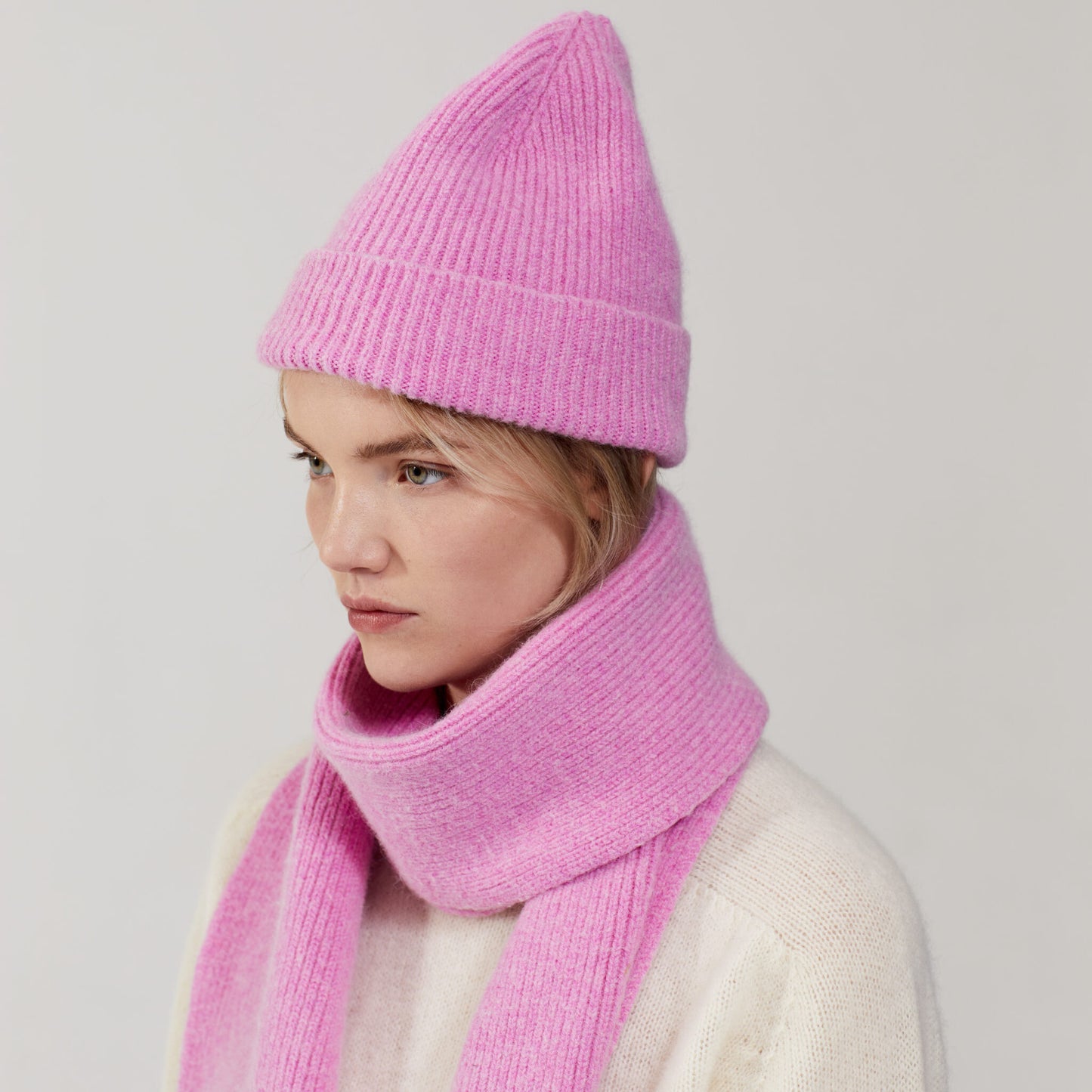 In a plain setting, a person wears Le Bonnet's pink knit hat, the Taffy scarf, and a cream sweater, glancing sideways.