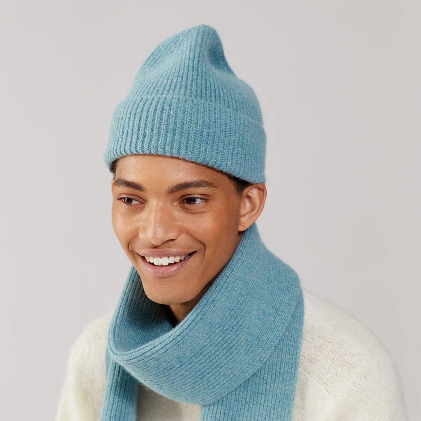 A person wearing a Basil beanie and matching scarf by Le Bonnet, smiling warmly in a neutral setting.