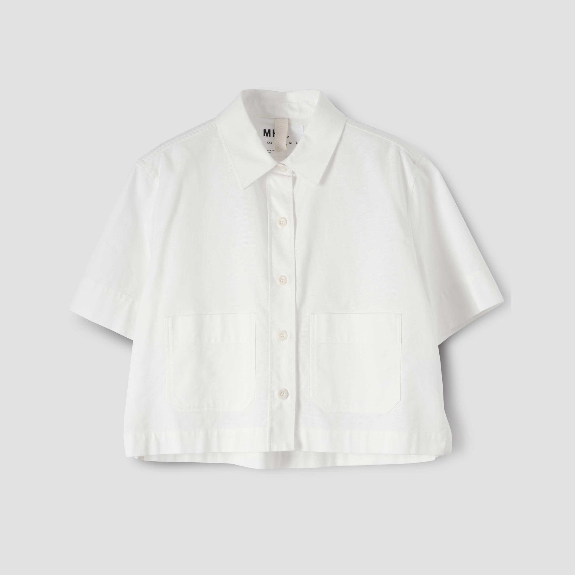 The W Cropped Uniform Shirt by Margaret Howell is a white, short-sleeve top with a Japanese panama weave and two chest pockets, shown on a plain grey background.