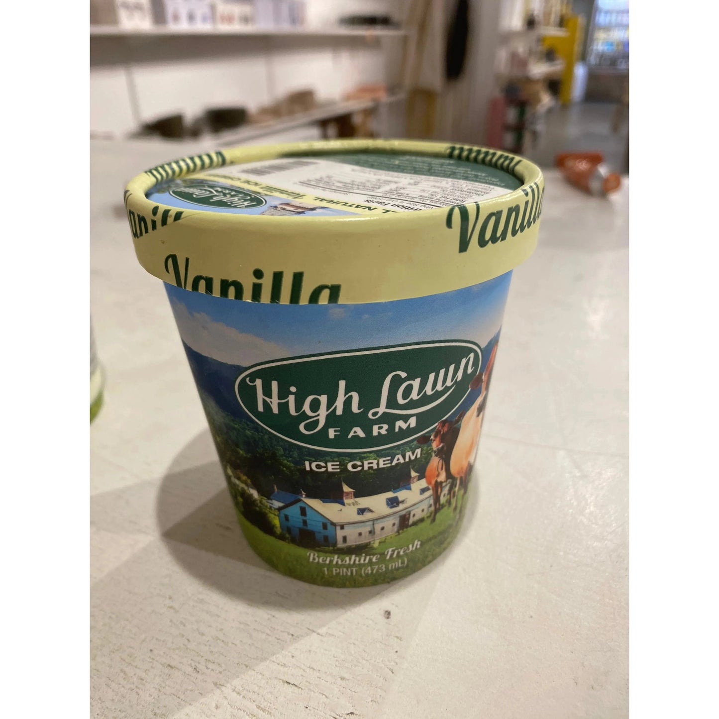 A pint container of High Lawn Farm's Vanilla Ice Cream - High Lawn Farm sits on a white surface, featuring a label with an image of a farm scene and a cow.