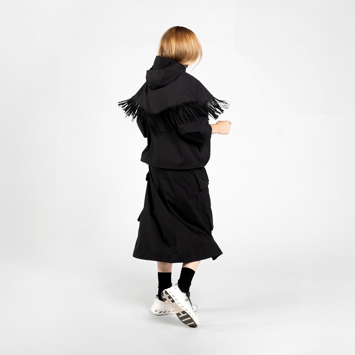 Person stands with back to camera, wearing an F/CE Pertex Water Repellent Fringe Hoodie, black skirt, black socks, and white sneakers against a plain white background.