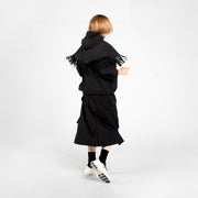 Person stands with back to camera, wearing an F/CE Pertex Water Repellent Fringe Hoodie, black skirt, black socks, and white sneakers against a plain white background.
