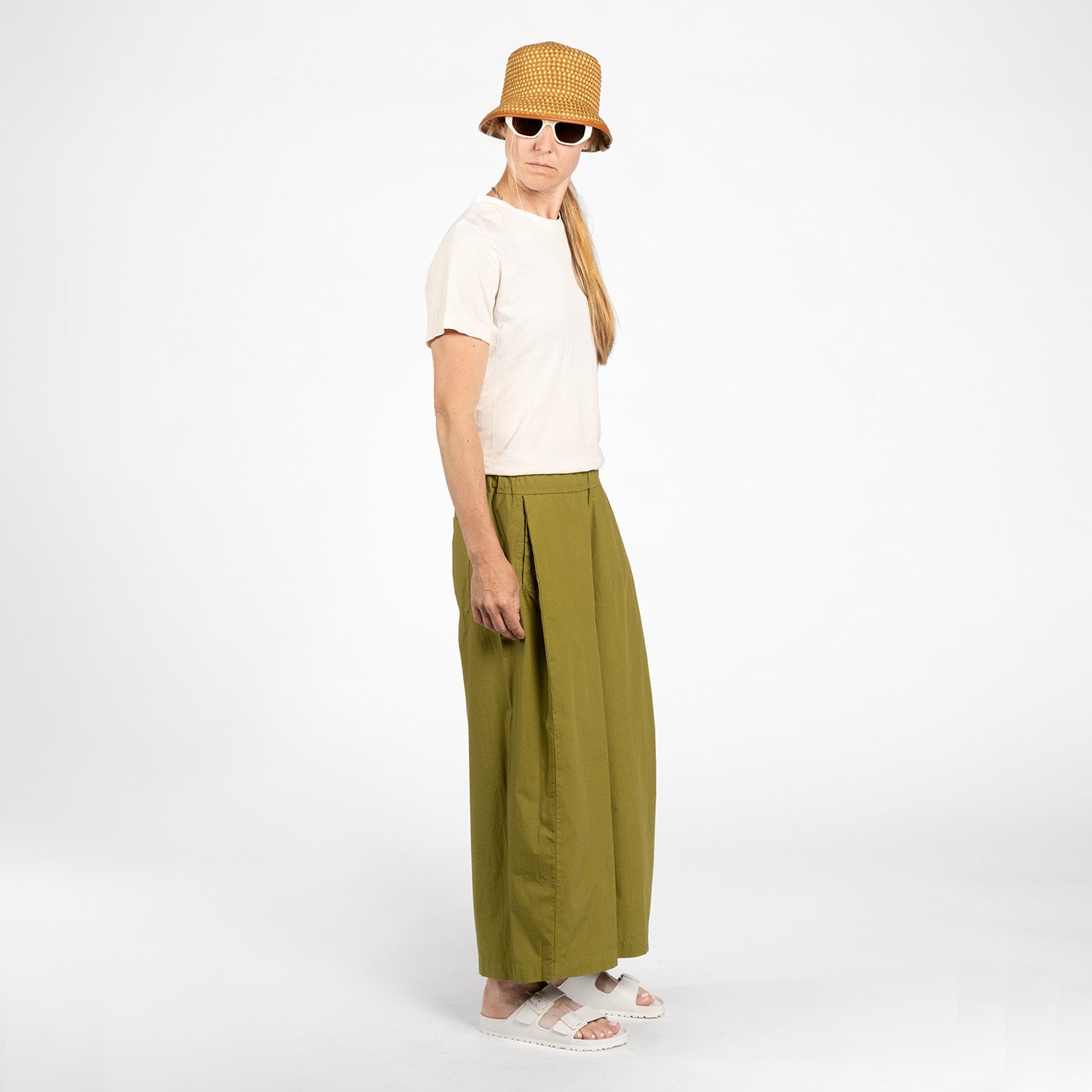 Model in beige t-shirt, Labo Art's Panta Storto Pant in Clara (olive green, high-waisted wide leg), white sandals, brown woven bucket hat, and white sunglasses stands against a plain white background.