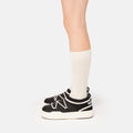 A person stands on a plain white background, sporting Moon Boot's sleek Park Icon slip-on sneakers in black and white, paired with high white socks. These sneakers boast an ultra-flexible construction.