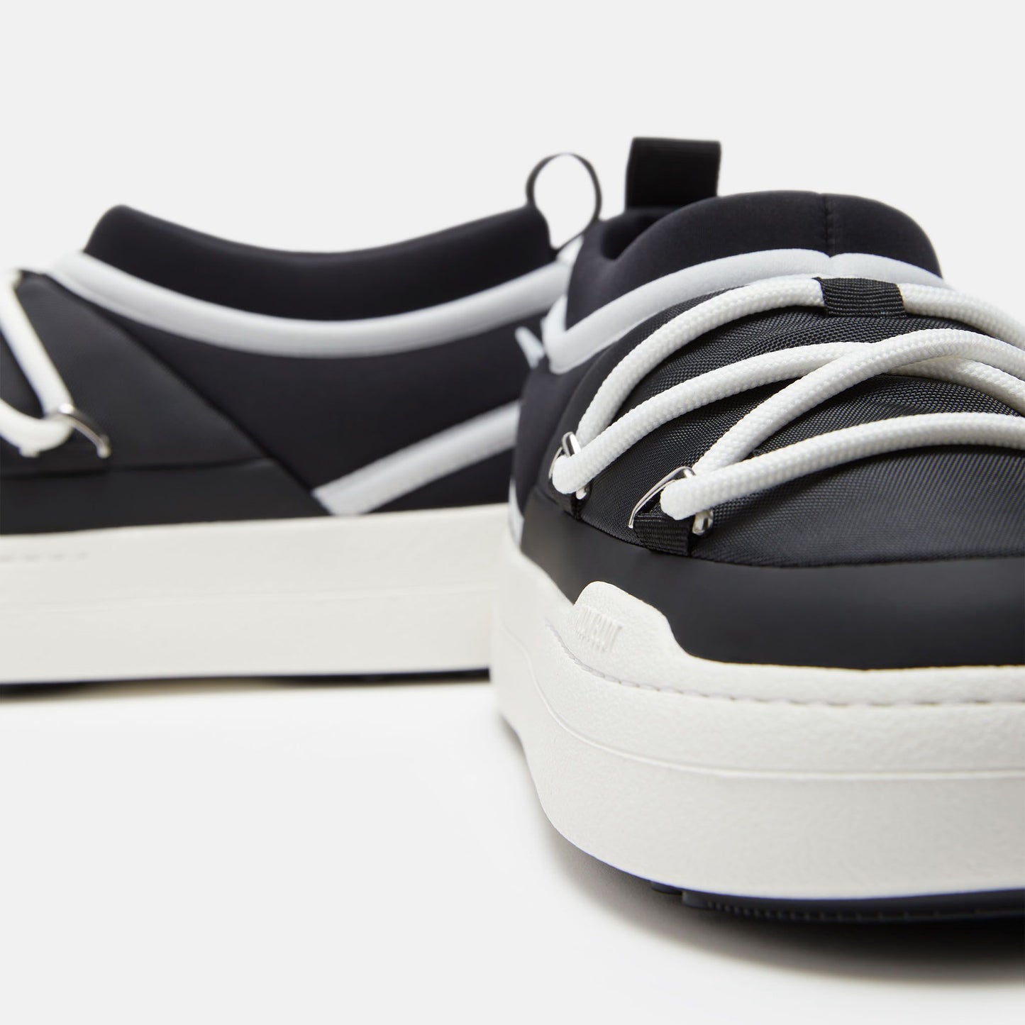 Close-up of Moon Boot's Park Icon sneakers, black and white with ultra-flexible construction, featuring thick white soles and intertwined laces set against a plain white background.