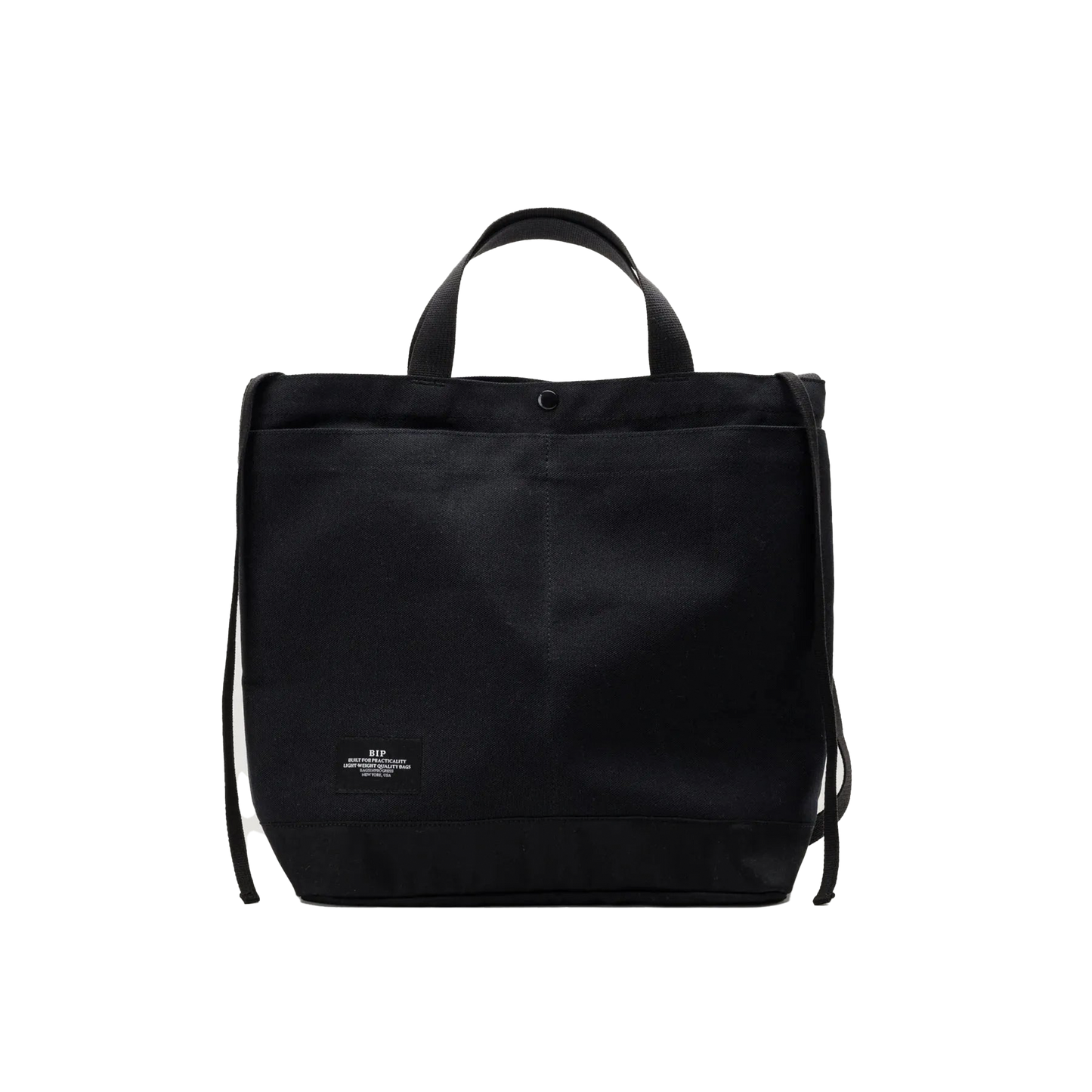 The Bags in Progress New York Tote Medium is a black rectangular cotton canvas bag made in Japan, featuring short handles, an adjustable shoulder strap, and a small white label at the bottom front center.