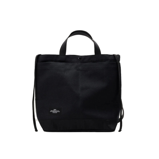 The Bags in Progress New York Tote Medium is a black rectangular cotton canvas bag made in Japan, featuring short handles, an adjustable shoulder strap, and a small white label at the bottom front center.