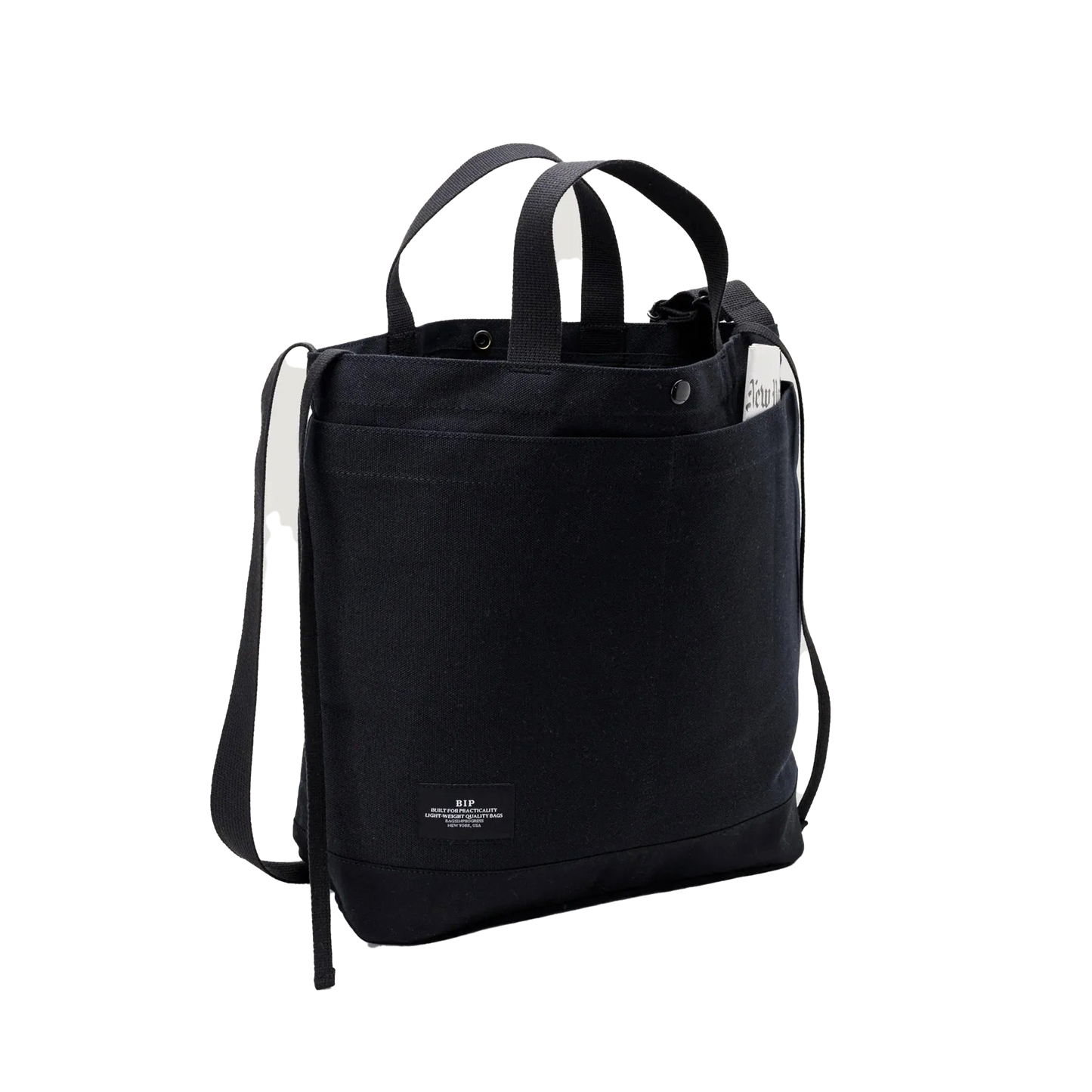 The Bags in Progress New York Tote Medium is a black cotton canvas bag made in Japan, with dual handles, an adjustable shoulder strap, a small white front label, and a document partially visible in the side pocket.