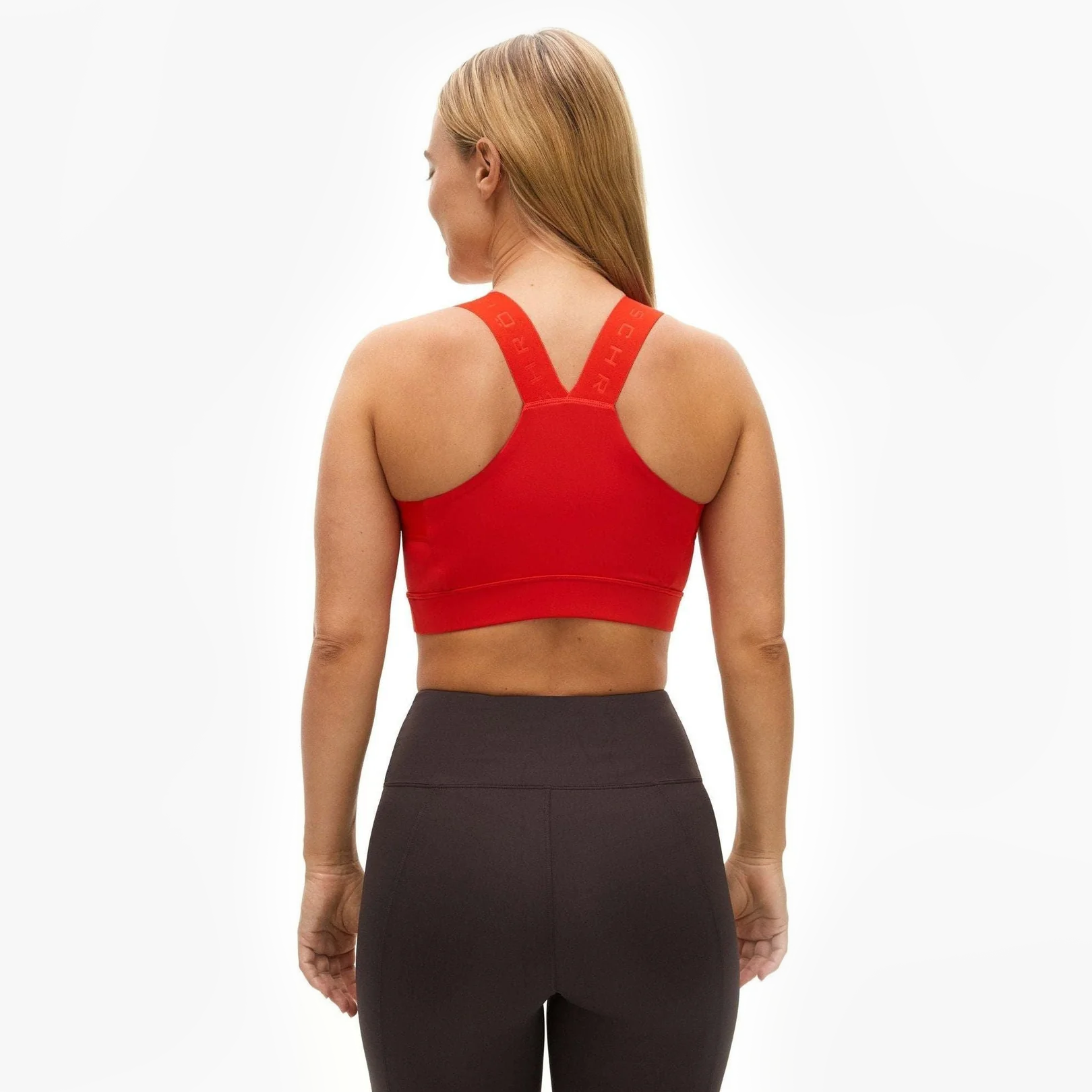 A woman faces away, wearing the Rohnisch Kay Sports Bra in red and high-waisted black leggings made from recycled polyester, set against a plain white background.