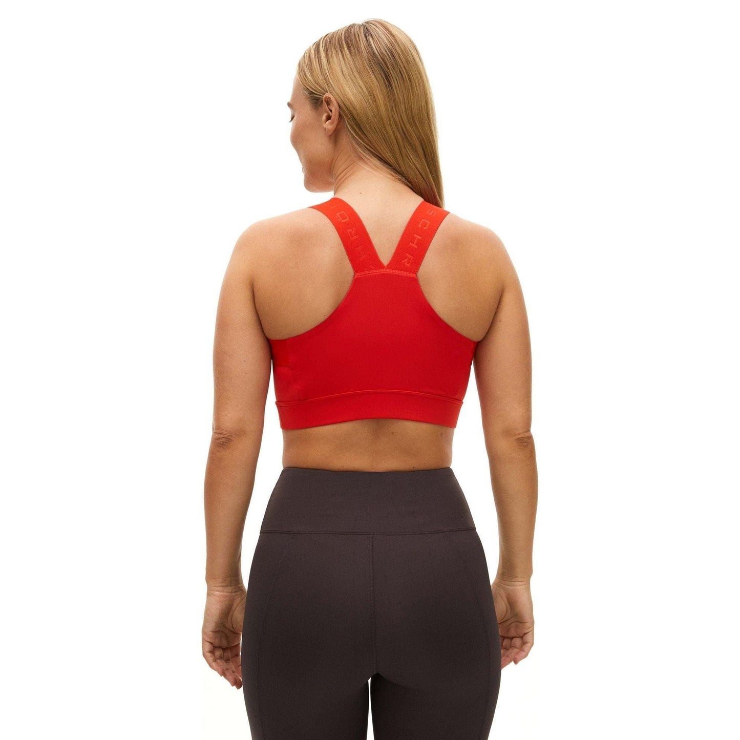 A woman, with her back to the camera, wears a Rohnisch Kay Sports Bra made from recycled polyester in red and black leggings. Her simple fitness attire contrasts against the plain white background, suggesting moisture-wicking comfort for workouts.