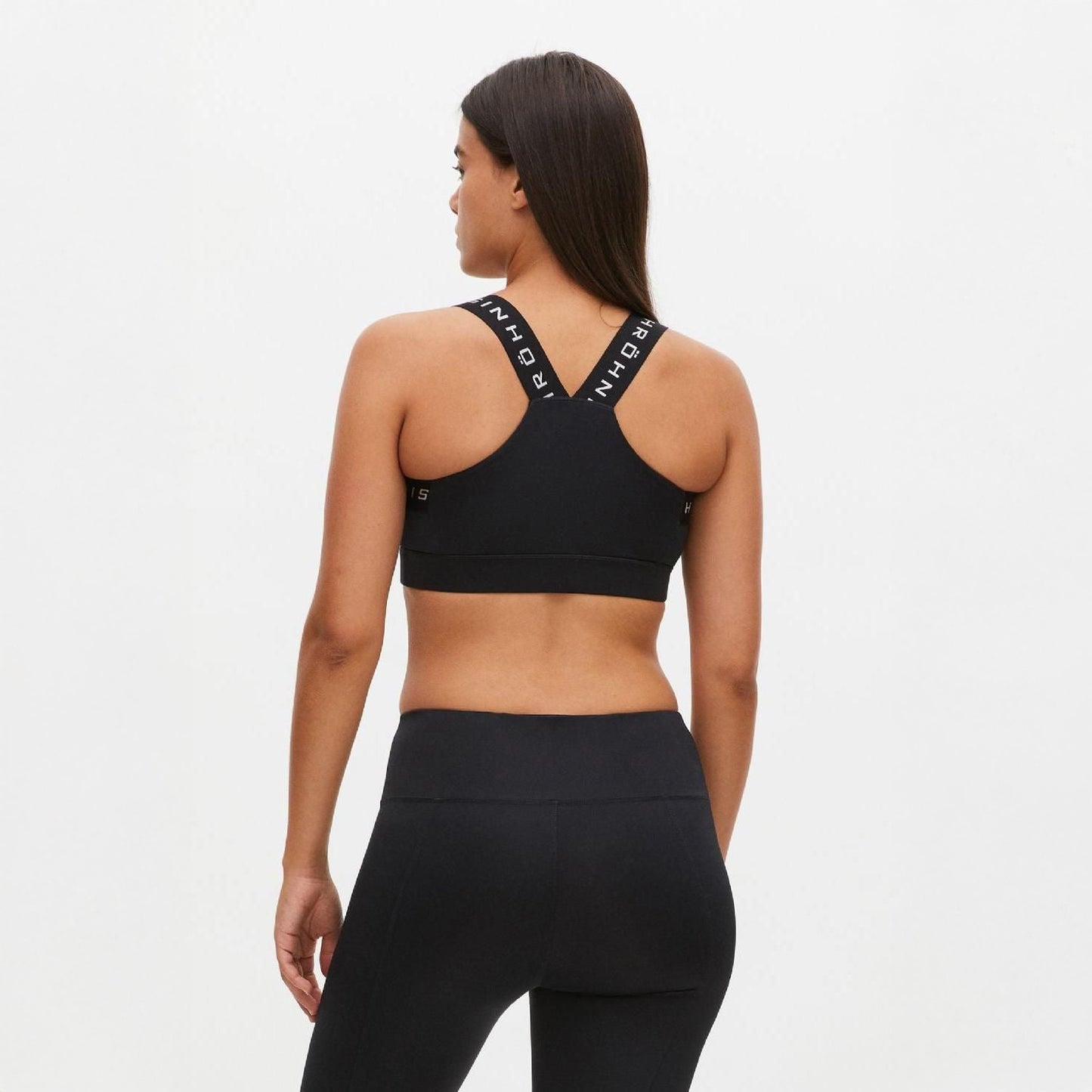 A woman models the back of Rohnisch's Kay Sports Bra with branded straps, paired with matching leggings in black athletic wear, all made from recycled polyester.