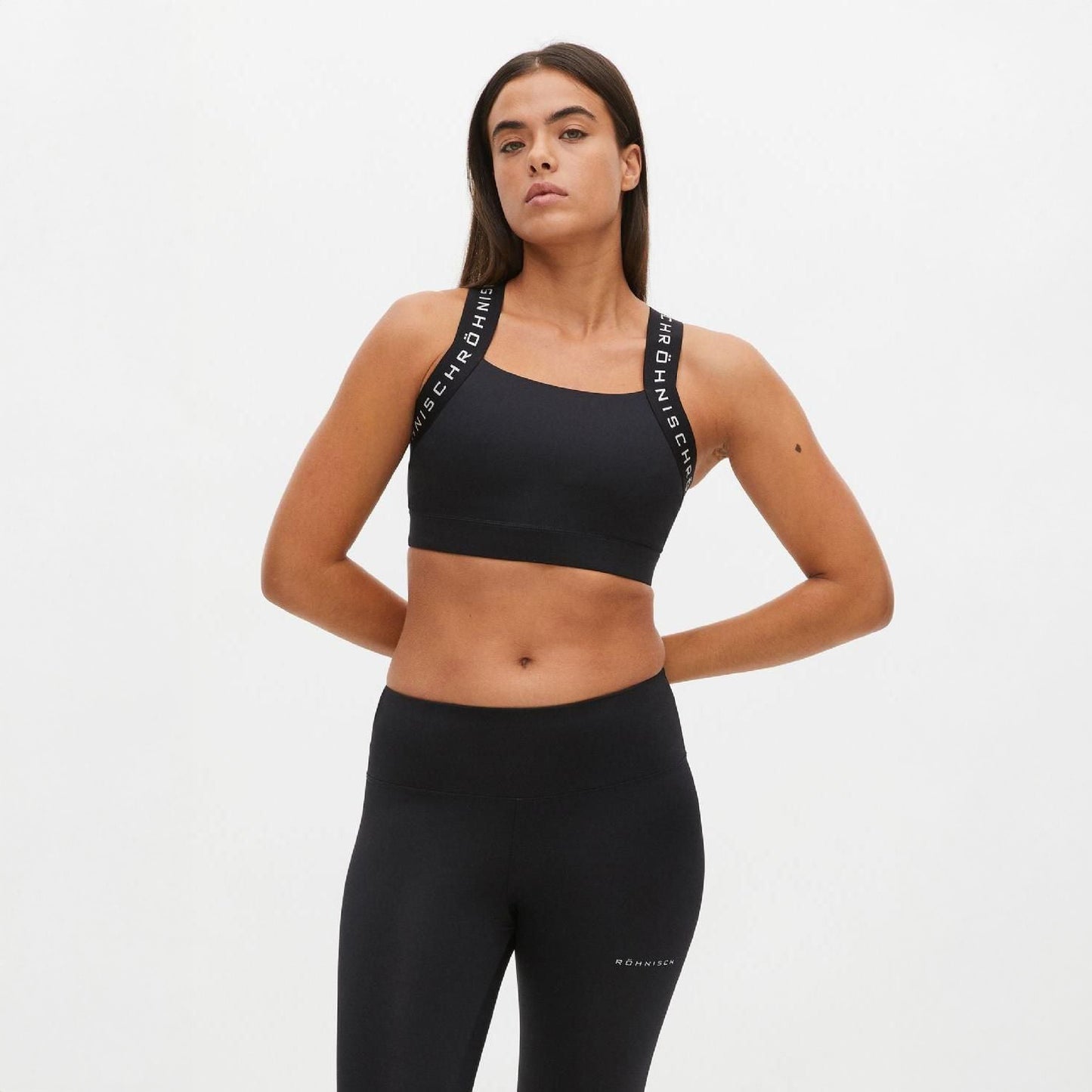 A person poses with hands on hips in black workout attire featuring the Rohnisch Kay Sports Bra, known for its breathable fabric and branded straps, set against a plain white background.